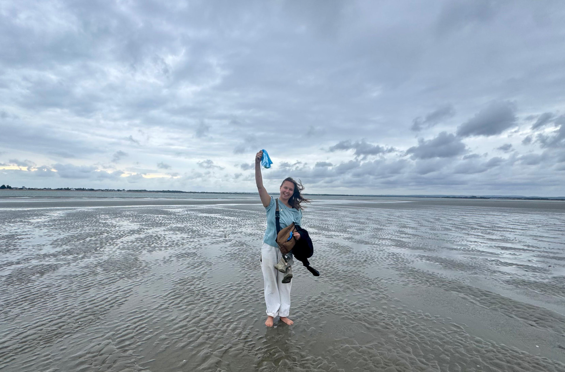 Phoebe Hyder standing barefoot on the wet sand flats at East Head, Sussex, holding up a piece of seaweed under a big cloudy sky